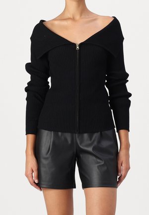 Femme portant un pull côtelé noir épaules dénudées avec une fermeture éclair sur le devant et un short en cuir noir sur un fond uni.