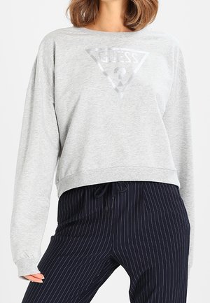 Woman wearing a light gray long-sleeve cropped sweatshirt with a faint triangular logo and navy pinstriped drawstring pants.