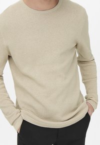 Beżowy dzianinowy sweter z fakturowanym wzorem, okrągłym kołnierzem i długimi rękawami, zestawiony z czarnymi spodniami. Tkanina wydaje się miękka i przewiewna.