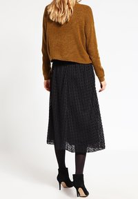 Brązowy sweter dzianinowy o luźnym kroju, zestawiony z czarną koronkową spódnicą. Czarne botki za kostkę dopełniają wygląd, ukazując projekt z warstwową teksturą.