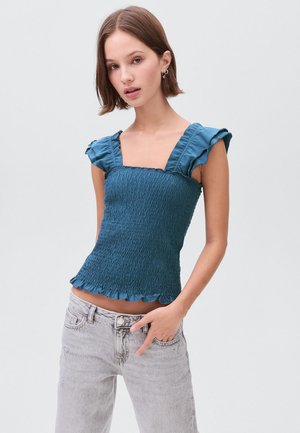 Teal smocked top with ruffled cap sleeves, square neckline, and a scalloped hem. Worn with light gray denim jeans.
