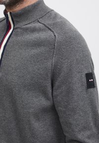Pulover gri tricotat cu fermoar, cu guler înalt, având o dunga tricolor (roșu, alb, albastru) de-a lungul fermoarului și o emblema pe mânecă.