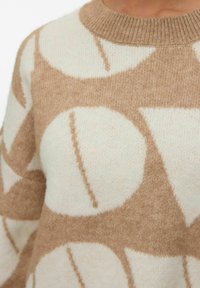 Pull beige et crème à motifs, présentant de grands motifs circulaires avec des lignes diagonales. Fabriqué en tissu tricoté doux et texturé.