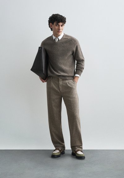 Maglione a maglia marrone, pantaloni marroni a quadretti e scarpe bicolore. Borsa tote nera in mano. Sfondo semplice, tonalità neutre.