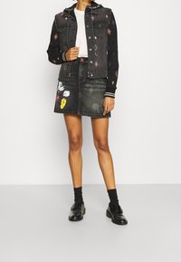 Person som bär en svart broderad denimjacka, vit skjorta, svart denimkjol med Mickey Mouse-motiv, svarta strumpor och svarta skor.