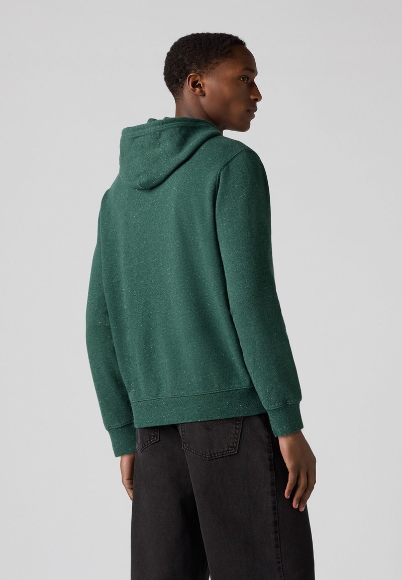 Grüne Kapuzenjacke aus strukturiertem Material, mit Kapuze, gerippten Bündchen und Saum, getragen mit dunklen Jeans, von hinten betrachtet.