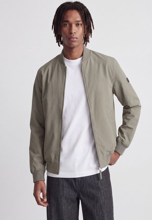 Jeune homme avec des dreadlocks portant une veste bomber vert olive clair, un t-shirt blanc et un pantalon foncé, debout devant un fond uni.