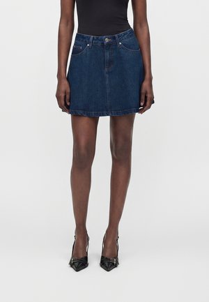 Femme portant une jupe en denim foncé, un top noir sans manches et des talons noirs pointus avec des détails de boucle, debout devant un fond uni.