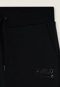 Pantalones jogger de algodón negros con una cinturilla acanalada. Presenta un logo de strass plateado en la parte delantera izquierda y bolsillos laterales. Textura suave.