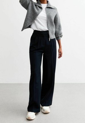 Personne portant un pull gris zippé, un t-shirt blanc, un pantalon noir à larges jambes et des baskets blanches, debout sur un sol gris clair.
