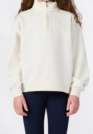 Persona che indossa una felpa bianca sporco con mezza cerniera e maniche lunghe, e pantaloni blu navy, in piedi davanti a uno sfondo semplice.