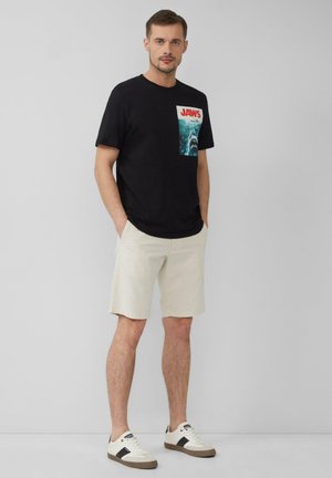 Zwart katoenen T-shirt met een Jaws-graphic op de borstzak, gecombineerd met lichtbeige shorts en witte sneakers met bruine accenten.