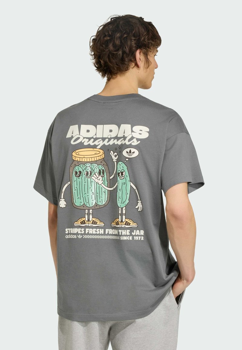Homme portant un t-shirt Adidas gris avec des cornichons dessinés, un dans un bocal, tous deux en baskets et faisant des signes de la main dans le dos.