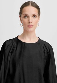 Blusa nera con scollatura rotonda, maniche corte a sbuffo e tessuto testurizzato. Il modello la abbina a orecchini a cerchio argentati.