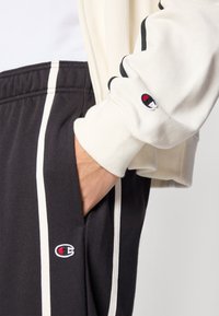 Μαύρα τζόγκερ με λευκές λωρίδες στο πλάι και πλαϊνές τσέπες, συνδυασμένα με μια κρεμ ζακέτα με φερμουάρ που έχει ένα μικρό κεντημένο λογότυπο στο μανίκι.