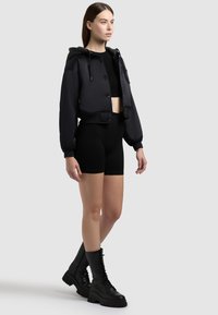 Jeune femme debout portant une veste à capuche noire raccourcie, un crop top noir, des shorts de cycliste noirs et des bottes de combat noires à lacets, sur un fond uni.