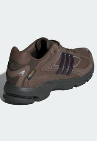 Brun idrottssko i mesh och mocka, med lila ränder, vadderad krage och gummisula med en synlig luftkudde.