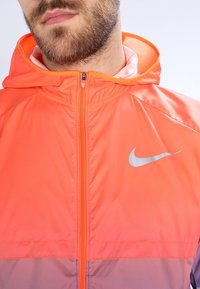 Chaqueta con capucha de Nike con diseño degradado en naranja brillante y púrpura, con un logo plateado en el pecho izquierdo y cierre de cremallera.