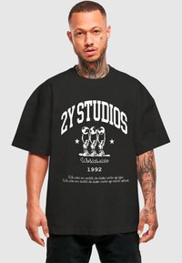Fekete, oversized T-shirt fehér szöveggel és grafikával. Tartalmazza a "2Y STUDIOS" és a "Worldwide 1992" feliratokat, valamint három figurát, amelyek hangsúlyozzák az elülsőt.