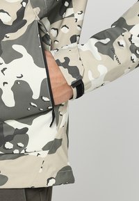 Veste camouflage présentant un mélange de motifs beige, gris et noir, avec une poche avant et un détail de réglage par Velcro au niveau des poignets.