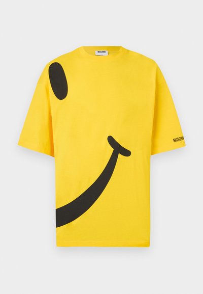 Κίτρινο βαμβακερό t-shirt με μεγάλο μαύρο γραφικό του χαμογελαστού προσώπου, φαρδιά γραμμή και λογότυπο στο μανίκι. Μαλακή υφή και κοντά μανίκια.
