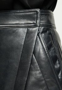 Κοντινό πλάνο μαύρης δερμάτινης φούστας με ραμμένες λεπτομέρειες πάνελ και φαρδιά ζώνη μέσης, φορεμένη πάνω από μαύρο ύφασμα.