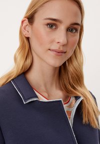 Blond naine heleda nahaga, seljas tumesinine jakk triibulise särgi peal, nägu pisut vasakule suunatud ja neutraalse ilmega.