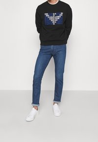 Sudadera negra con el logo "Emporio Armani" en azul y blanco, combinada con jeans oscuros enrollados y zapatillas blancas. Diseño simple y ajustado.