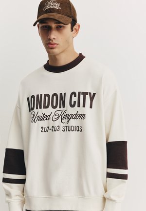 Bluză sport crem cu dungi orizontale maro închis pe mâneci. Are inscripția „LONDON CITY” și „United Kingdom” în text maro.