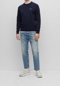 Sudadera de cuello redondo navy con detalle de logo, combinada con jeans claros desgastados y zapatillas beige. Un atuendo casual y en capas.