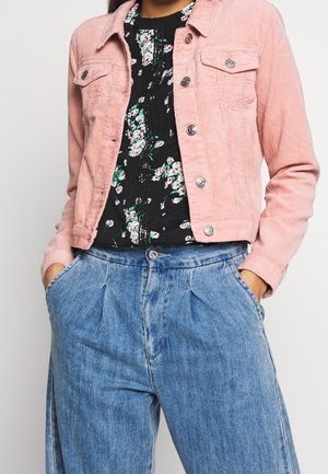 Osoba w różowej krótkiej kurtce sztruksowej, czarnej kwiecistej bluzce i wysokich plisowanych niebieskich dżinsach z rękami w kieszeniach.