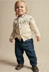 Ljust beige stickad cardigan med blå blommönster, matchad med marinblå byxor och grå skor. Barnet står på en neutral bakgrund.