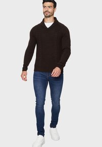 Mørkebrun strikket sweater med sjalskrave, prydet med et struktureret mønster og ribbede manchetter, parret med tætsiddende blå jeans og hvide sneakers.
