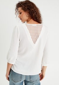 Haut blanc avec un dos dentelé en forme de V, des manches longues bouffantes et un tissu texturé. Associé à un jean bleu clair.
