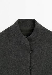 Manteau en laine gris foncé avec un col haut et arrondi, et deux boutons noirs visibles sur le devant.