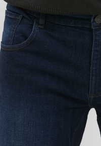 Jeans de denim azul oscuro con un ligero estiramiento, que cuentan con un bolsillo frontal y costuras visibles. La tela tiene una textura suave con un corte clásico.