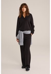 Femeie cu păr ondulat purtând un top negru cu mâneci lungi și pantaloni, cu o cămașă în dungi alb-negru legată în jurul taliei, stând într-o poziție neutră.