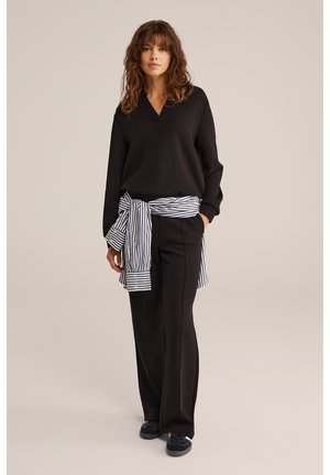 Naine laineliste juustega, musta pikkade varrukatega topi ja pükstega, must-valge triibulise särgi vööl seotud, seisab neutraalselt.