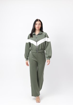 Kvinde iført olivengrønt træningssæt med hvidt chevronstribe, stående mod en hvid baggrund, vendt fremad med neutralt udtryk.