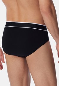 Slip neri in cotone con una fascia elasticizzata a righe bianche, design aderente, texture liscia e taglio a vita media, mostrano una vista laterale e dettagli minimali.