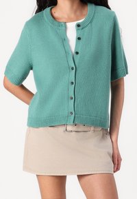 Krátký rukáv cardigan v teplé modré pletenině, s kulatým výstřihem, zapínáním na knoflíky a žebrovanými detaily na rukávech a lemu. Nosený přes bílý top.