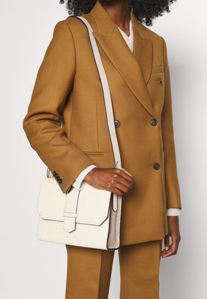 Mujer con traje marrón de doble abotonadura que sostiene un bolso de cuero crema con solapa y detalle de hebilla.