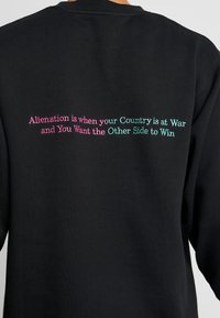Felpa nera con testo ricamato in rosa e teal sulla parte posteriore che recita: "L'alienazione è quando il tuo Paese è in guerra e tu desideri che l'altra parte vinca."