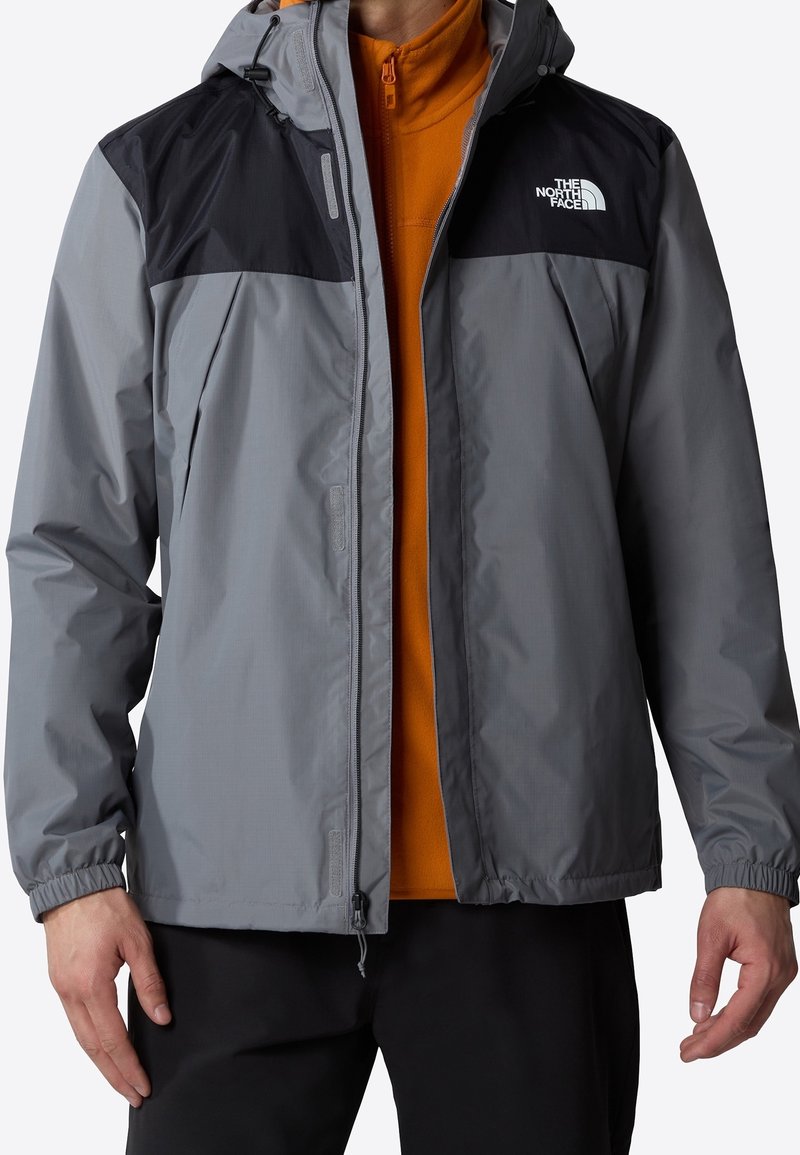 Homme portant une veste imperméable North Face grise et noire par-dessus une polaire orange avec un pantalon noir, debout, bras relâchés.