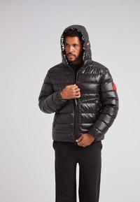 Giacca nera in piuma con finitura lucida, chiusura frontale con zip e cappuccio regolabile. Presenta una toppa rossa e grandi tasche laterali.
