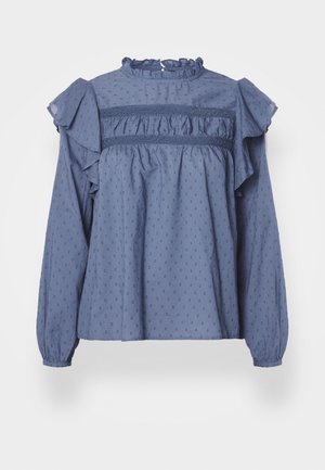 Blue cotton blouse with a ruffled neckline and shoulders, featuring a textured dot pattern and gathered detailing along the yoke and sleeves.