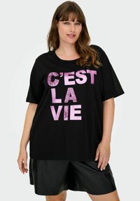 Camiseta de manga corta negra con "C'EST LA VIE" en diferentes tonos de rosa, con un corte holgado y hecha de material de algodón suave. Combinada con pantalones cortos negros.