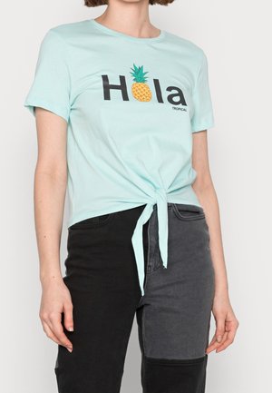 Kobieta ubrana w jasnoniebieską koszulkę z wiązaniem z przodu, z napisem "Hola" i grafiką ananasa, w połączeniu z dżinsami dwukolorowymi w odcieniach czerni i szarości.