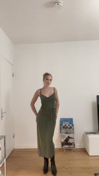 Olīvzaļš bezpiedurkņu kleita ar pieguļošu augumu, sasienamu vidukli un potītes garuma svārkiem, apvienota ar melniem potīšu zābakiem.