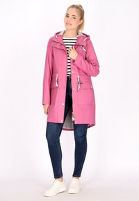 Giacca impermeabile rosa con fodera interna grigia, tasche frontali, cappuccio regolabile e stringhe. Abbinata a jeans scuri e sneakers bianche.